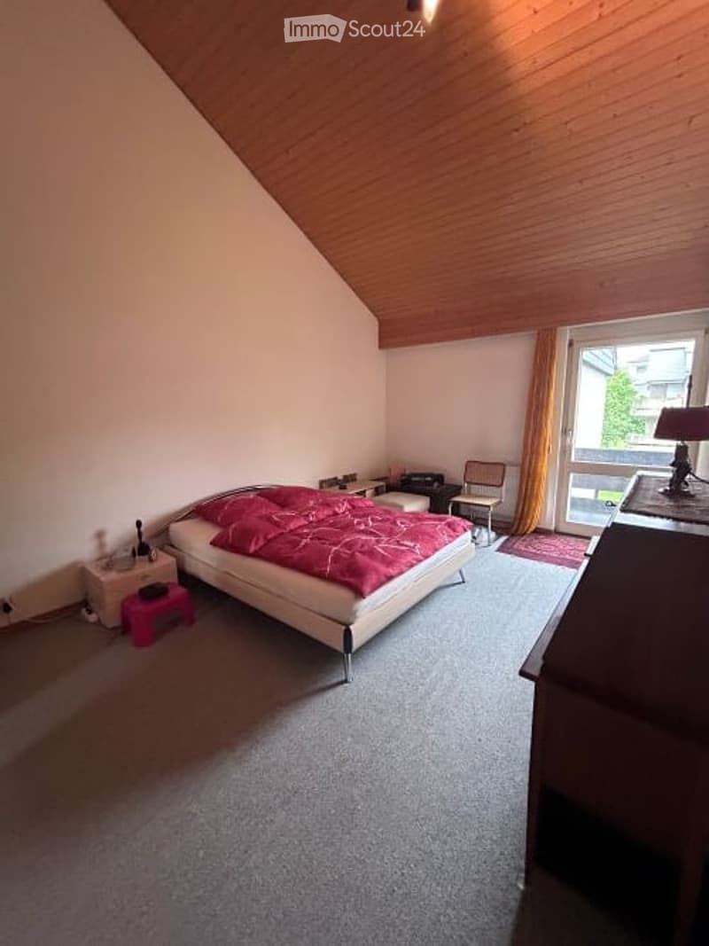 Cozy bedroom with a slanted wooden ceiling, grey carpet, a large bed with red bedding, nightstand, small stool, chair, and a desk with a lamp.