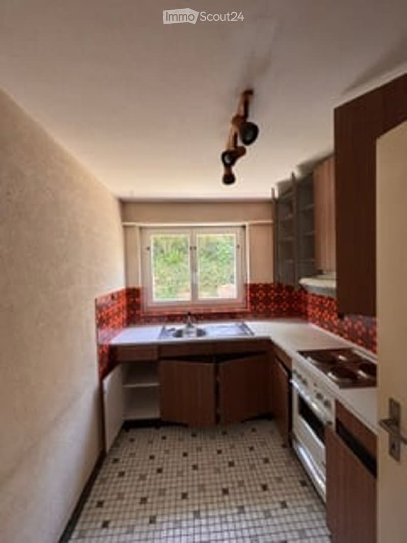 A small kitchen with wooden cabinets, a sink, a stove, and a window. The floor is tiled, and the walls have a red backsplash.