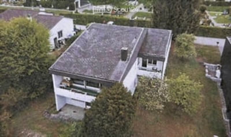 A two-story house with a grey roof and white walls, surrounded by greenery and trees. The house has multiple windows and a small balcony on the upper floor.