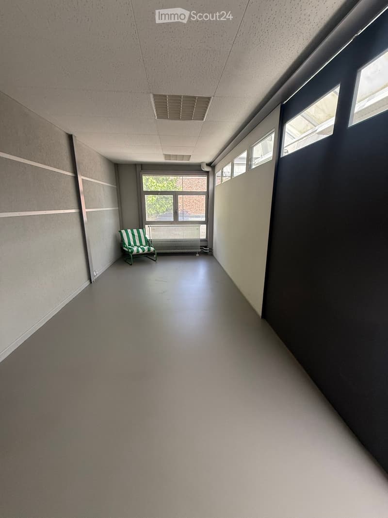 A long, narrow hallway with grey walls, carpeted floor, and a single green and white striped chair placed against the left wall. Large windows at the far end provide natural light, and the ceiling features recessed lighting and ventilation grilles.