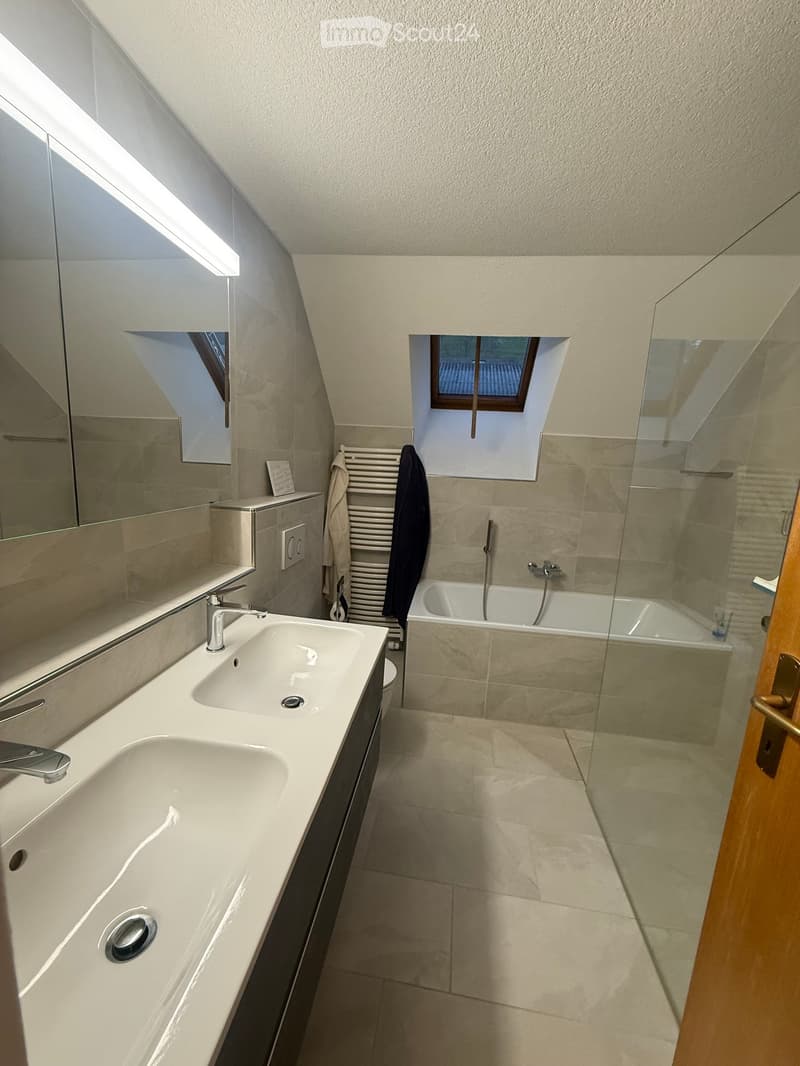 Modern bathroom with a double sink, bathtub, large mirror, radiator, and towel hanging on it. The room features a skylight window and a glass-enclosed shower area.
