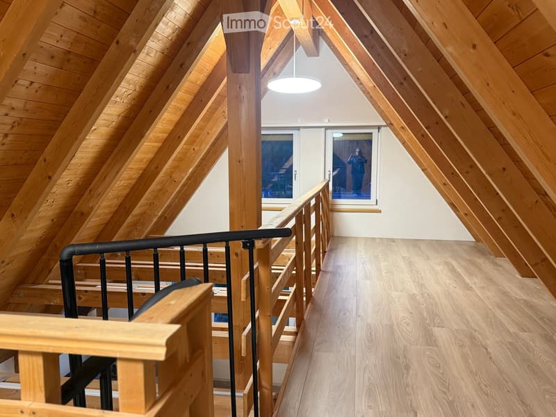 Bright, wooden-floored hallway with slanted wooden ceiling beams, a modern black railing, and a large window at the end.