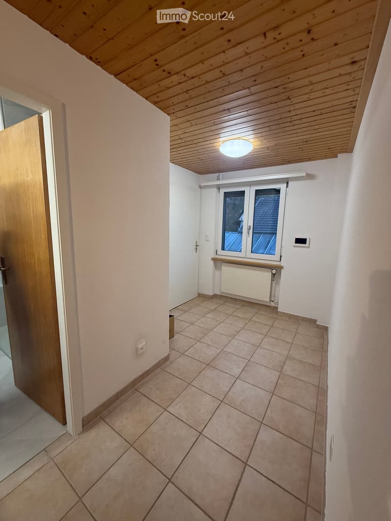 Empty hallway with tiled floor, white walls, wooden door, and a window with a view of the roof.