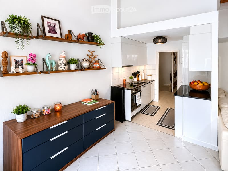 Modern kitchen with white cabinets, wooden countertops, and various decorative items on shelves. Includes an oven, fridge, and a small seating area with a table and chair.