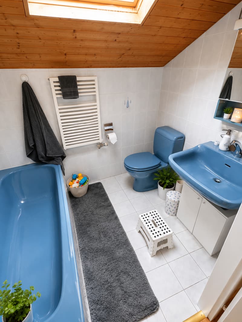 Modern bathroom with blue fixtures, including a bathtub, toilet, and sink. Features a towel rack, a small stool, a bath mat, and decorative plants. The room has white tiled walls and a skylight window.