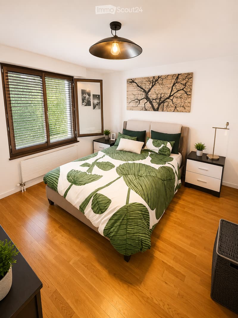 Modern bedroom with wooden flooring, large window with blinds, bed with green and white bedding, nightstands, lamp, mirror, and artwork on the wall.
