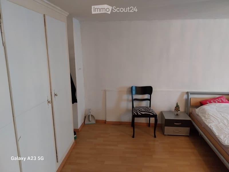 A small, furnished bedroom with a bed, a nightstand, a chair, a small plant, and a wardrobe. The room has light-colored walls and wooden flooring.