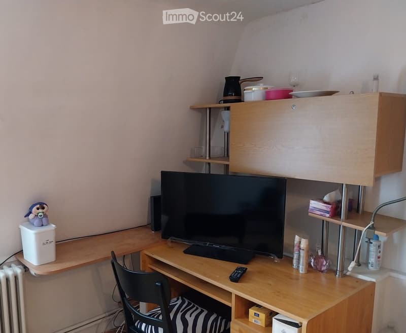 A small, furnished room with a wooden table, a black chair, a large screen, a remote control, a small cabinet, and various items on the shelves and table.
