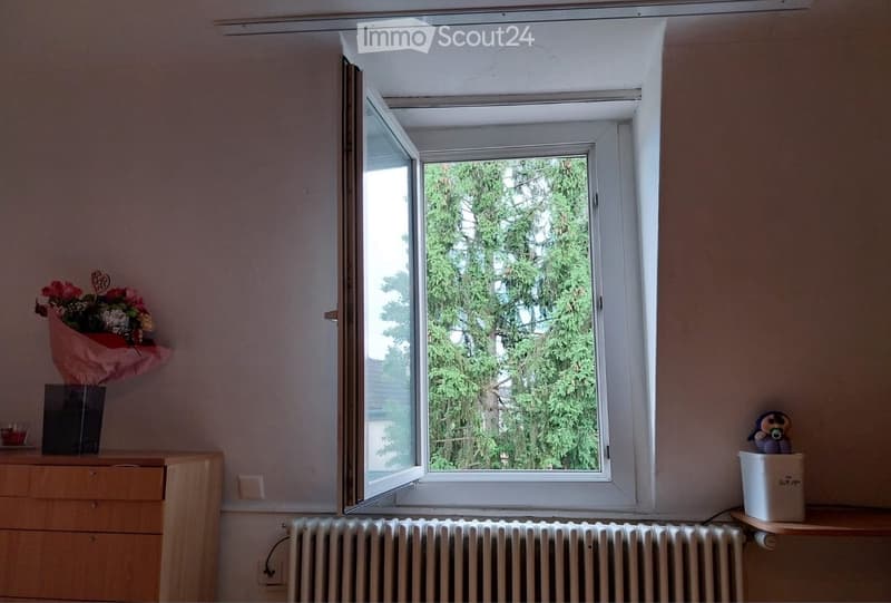 A small room with a large window showing greenery outside. The room features a wooden cabinet with a flower arrangement, a radiator below the window, and a small toy on a shelf.