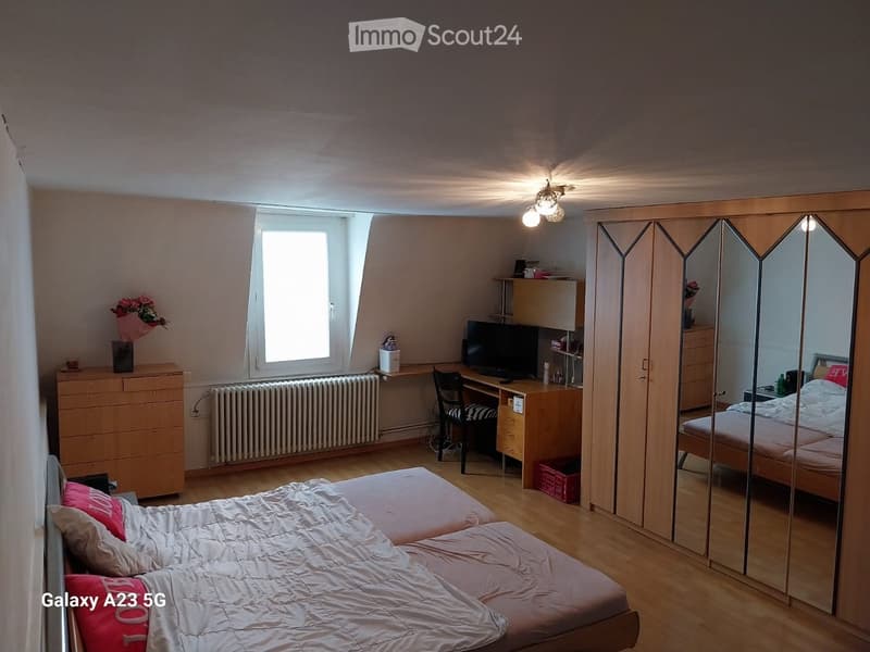 A well-lit bedroom with a double bed, a large mirror, a desk with a chair, and a window letting in natural light.