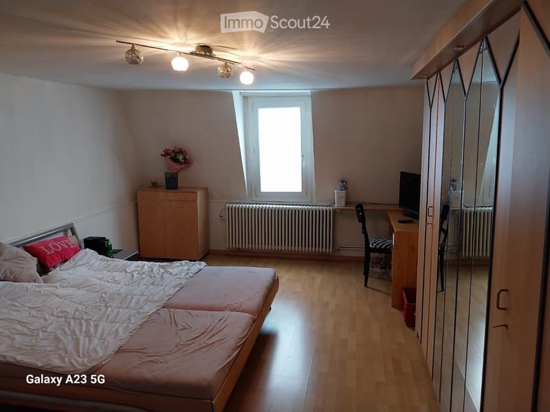 Cozy bedroom with a double bed, wooden floor, large window, desk with chair, mirror, and a small flower vase on a drawer unit.