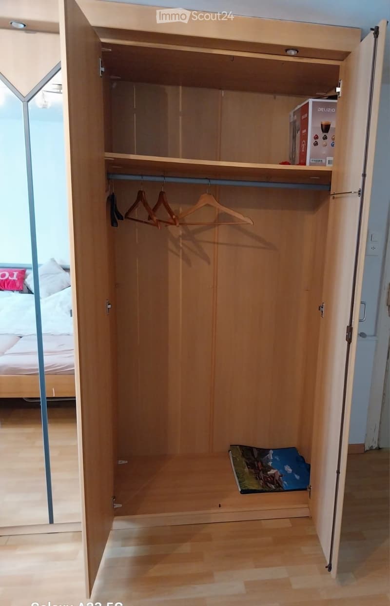 Open wooden wardrobe with shelves, hanging space, and a few stored items including a box and folded clothes.