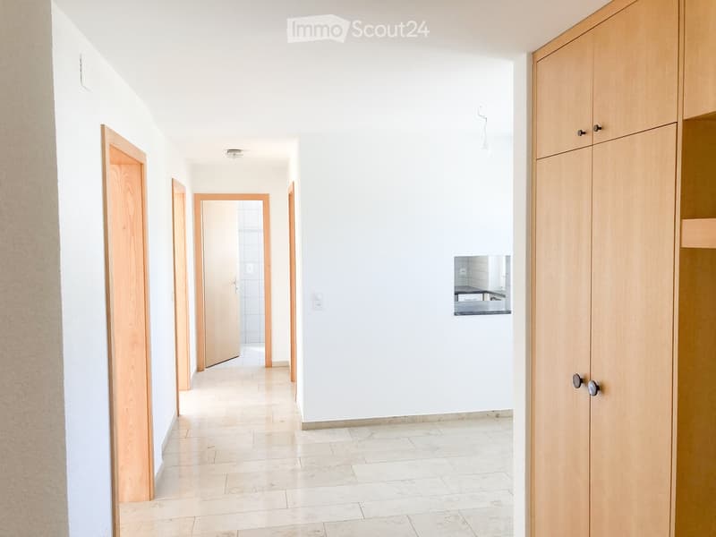 Empty hallway with white walls, tiled floor, and wooden doors leading to other rooms. A small mirror on the wall shows a glimpse of a kitchen area.