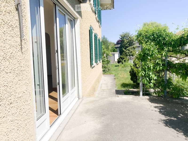 A concrete pathway leads to a house with green shutters and a textured beige facade. The area is surrounded by lush greenery and trees, creating a serene outdoor setting.