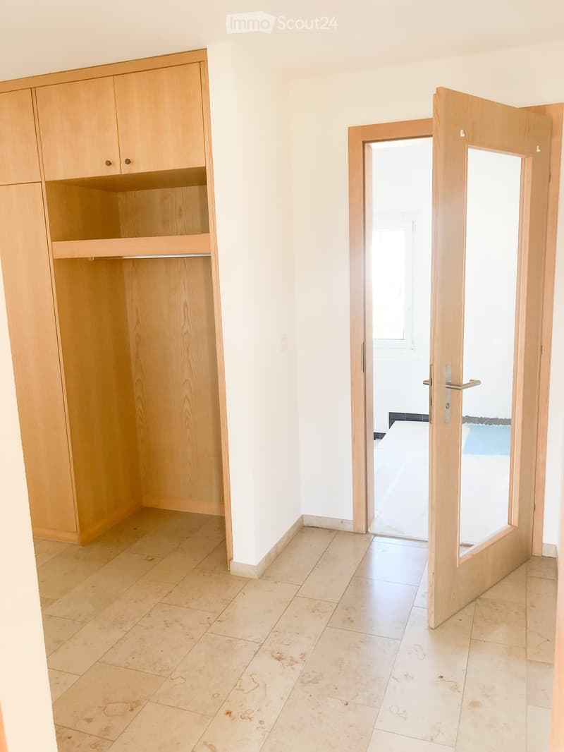 Empty room with light wooden built-in closet and an open doorway leading to another space. Light-colored tiled floor and white walls.