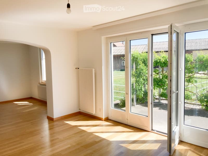 Empty room with wooden flooring, white walls, large sliding glass doors leading to a garden, and radiators on the walls.