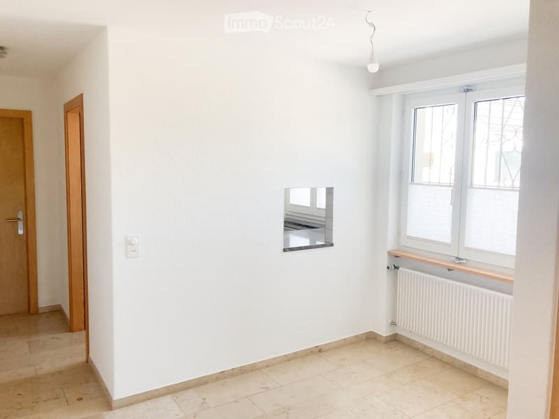 Empty room with white walls, tiled floor, two wooden doors, a window with a radiator, and a small mirror on the wall.