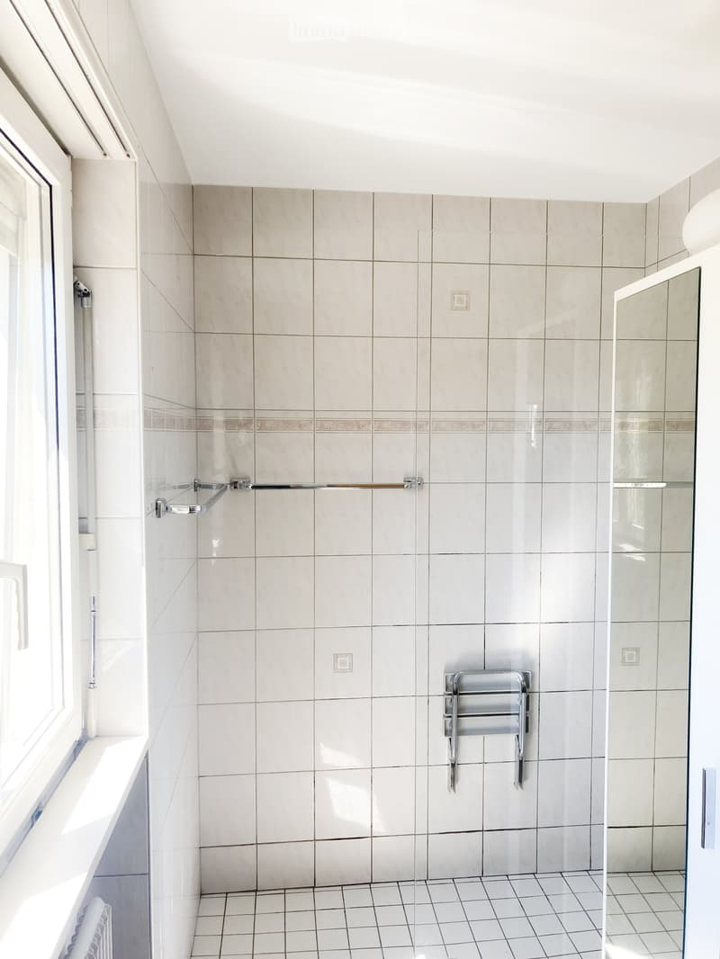 Modern bathroom with beige tiled walls, white tiled floor, glass-enclosed shower stall, towel rack, and a window with a white frame.