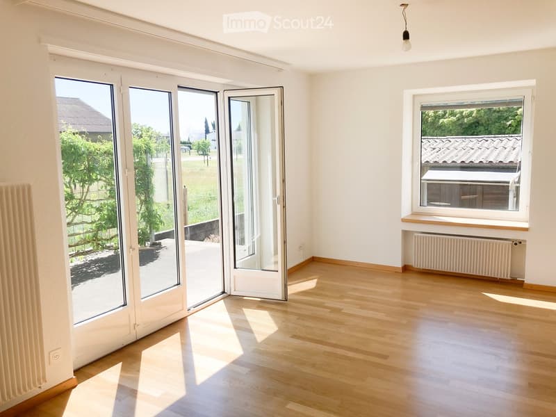 Empty room with wooden flooring, large sliding glass doors, and a window with a radiator below. Natural light fills the space.