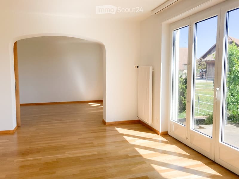 Empty room with light wooden flooring, white walls, and large windows letting in natural light. There is a radiator on one wall and a doorway leading to another space.
