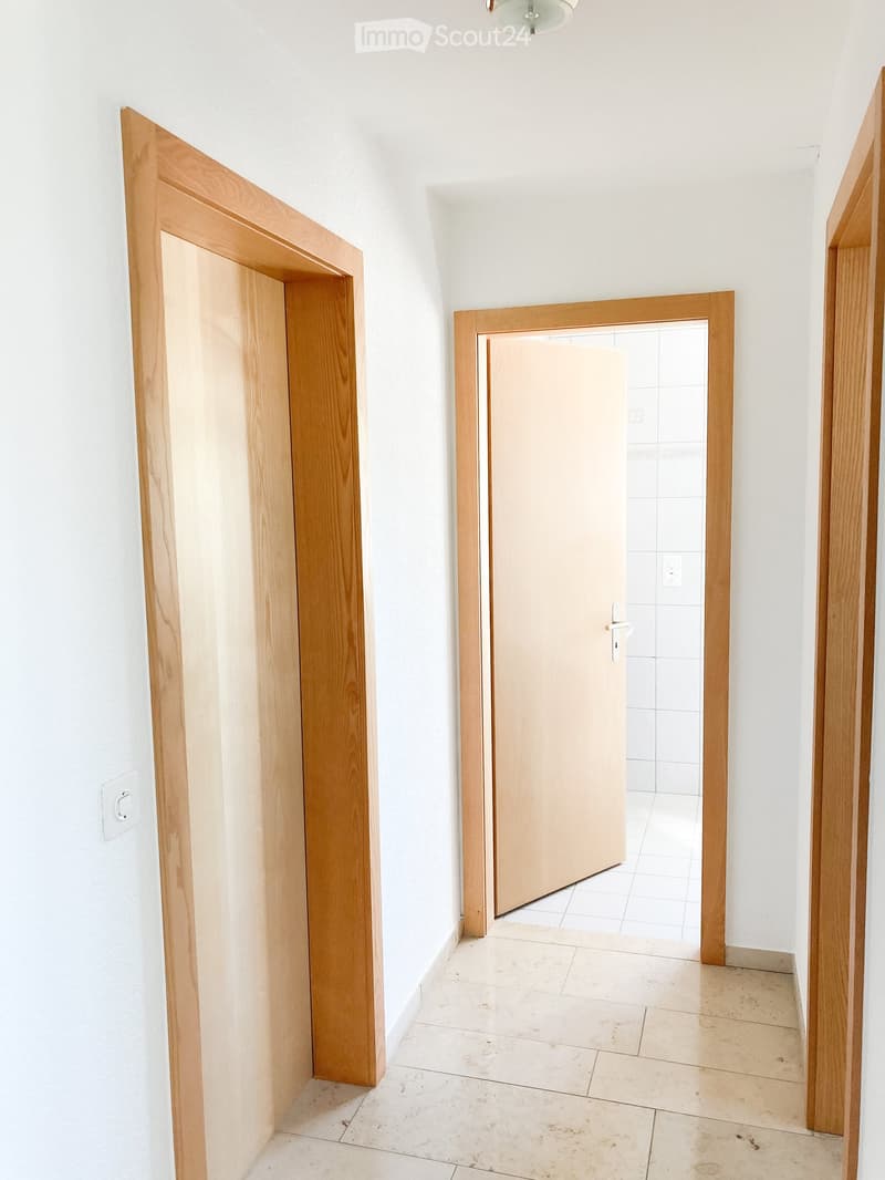 Empty hallway with light wooden doors and white tiled floor, leading to another room with tiled walls.