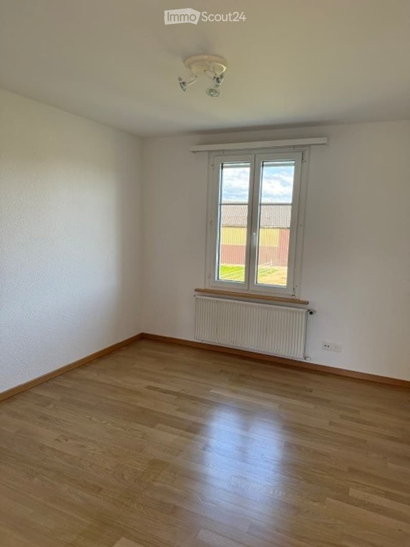 Empty room with wooden flooring, white walls, a window with white frame, radiator, and a ceiling light fixture.