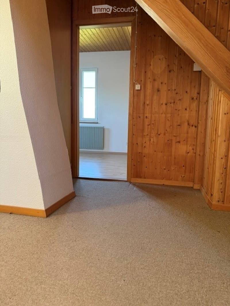 Empty room with wooden walls, carpeted floor, and a small window in the adjacent room.