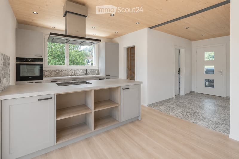 Modern kitchen with light grey cabinets, a central island, built-in oven, range hood, and a sink area. The space features wooden flooring and a mix of open shelving and closed storage.