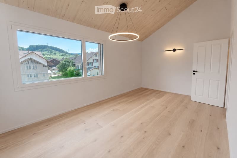 Empty room with light wooden flooring, white walls, large windows offering a scenic view, and modern lighting fixtures.