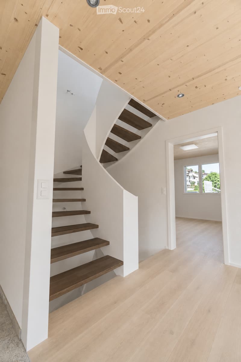 Modern hallway with wooden staircase, light wooden flooring, white walls, and exposed wooden ceiling beams.