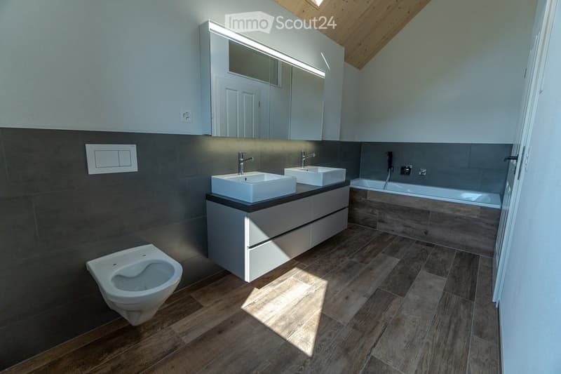 Modern design bathroom with wooden floors, gray tiled walls, two sinks with built-in drawers, a bathtub, a toilet, and an overhead mirror with lighting.