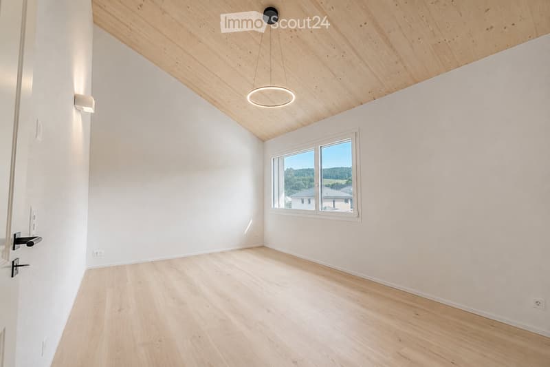 Empty room with light wooden flooring, white walls, and a large window offering a view of the landscape. The ceiling features exposed wooden beams and a modern pendant light fixture.