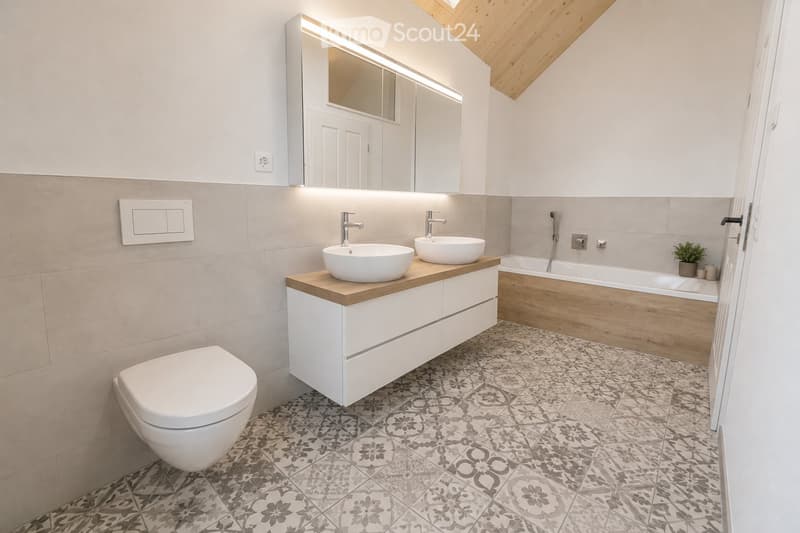 Modern bathroom with a white toilet, double sink vanity with wooden countertop, bathtub, large mirror with LED lighting, and decorative patterned tiles.