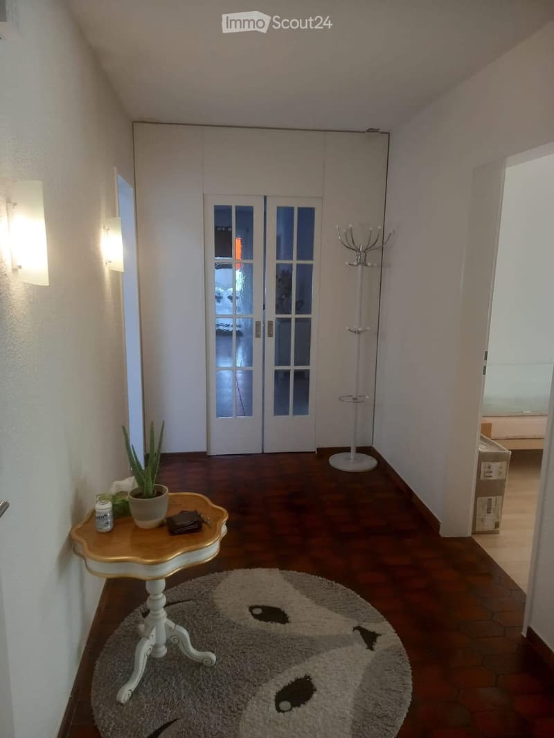 A hallway with a wooden floor, a round table with a potted plant, a coat rack, and a white wall with a light fixture.