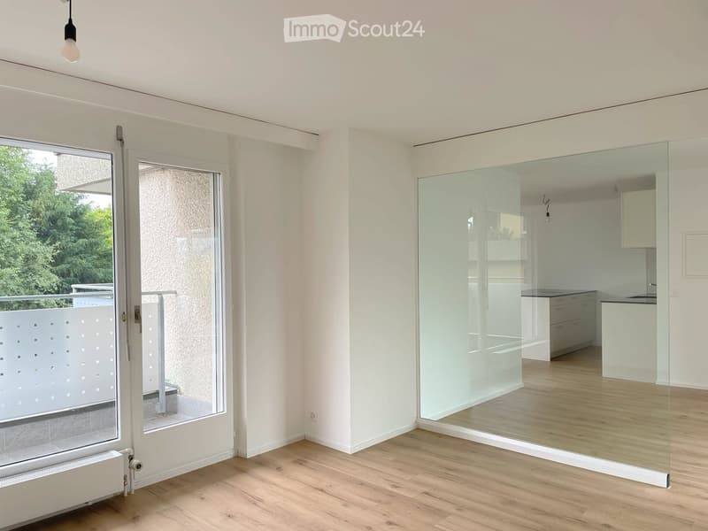 Bright, empty room with wooden flooring, white walls, large glass door leading to a balcony, and a glass partition separating the space from a kitchen area.