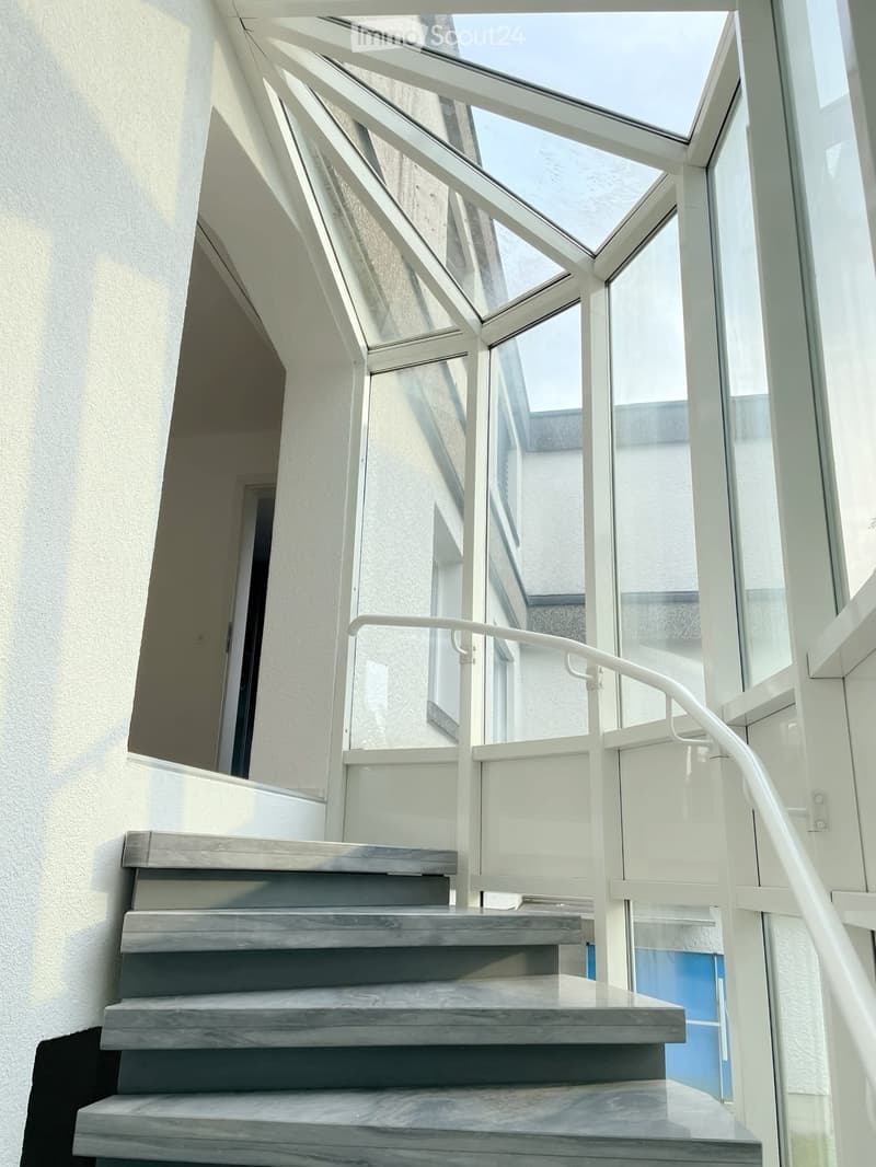 Modern staircase with wooden steps and white glass railing, leading to a bright, open area with large windows and a skylight above.