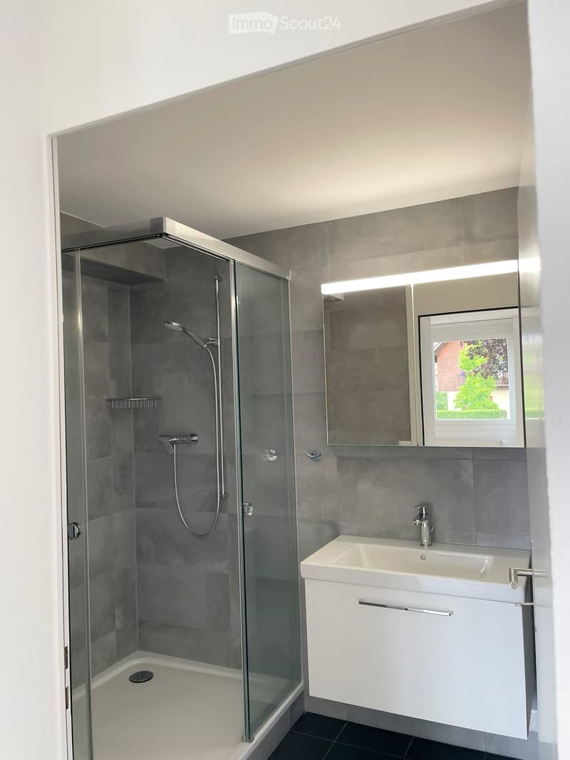 Modern bathroom with a glass-enclosed shower, white sink, and large mirror with built-in lighting. Grey tiles and black flooring.