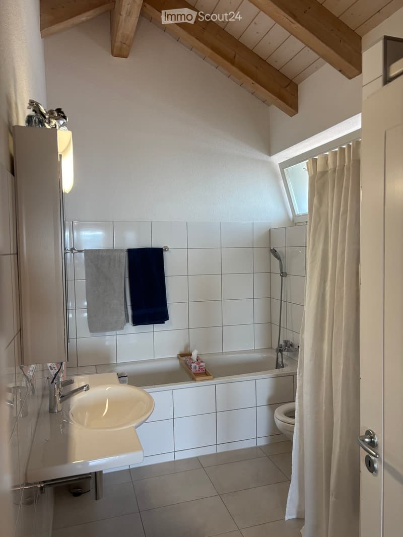 Bathroom with white tiled walls, a sink, bathtub, toilet, and a window with a curtain. Wooden beams on the ceiling and a small shelf with a tissue box are visible.