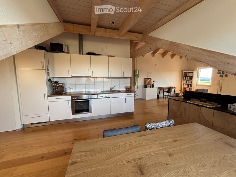 Open-plan living and dining area with a wooden table, chairs, sofa, bookshelf, desk, and a modern kitchen featuring white cabinets, stainless steel appliances, and a microwave.