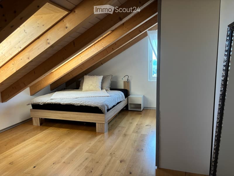 Cozy attic bedroom with wooden slanted ceilings, large window, wooden floor, bed with white bedding, nightstand, and lamp.