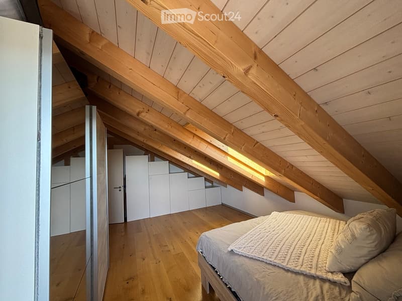Attic bedroom with wooden beams, slanted ceiling, wooden floor, bed with white bedding, white cabinets, and built-in storage.