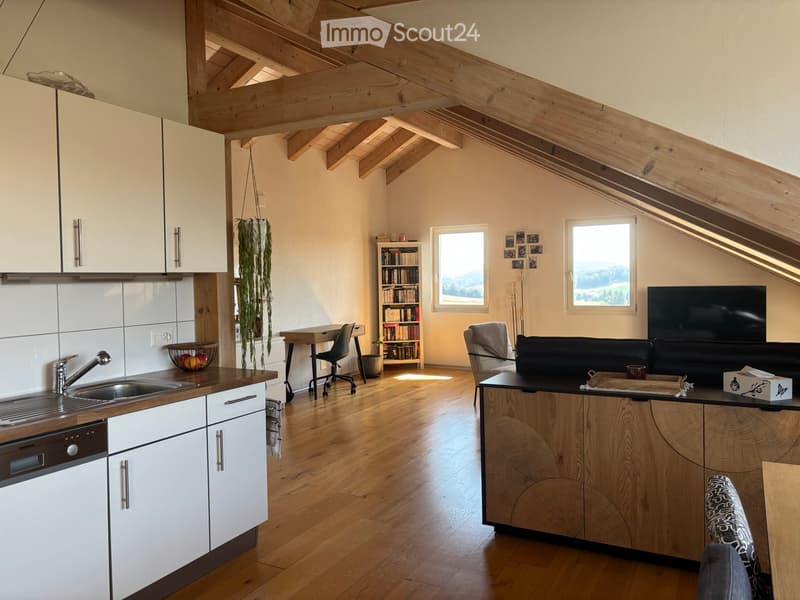 Open-plan living and kitchen area with exposed wooden beams, hardwood flooring, and large windows. Features a sofa, armchair, coffee table, bookshelf, sink, and modern kitchen cabinets.