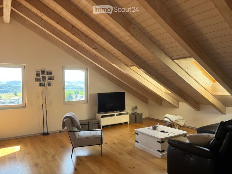 Bright living room with wooden floors, slanted wooden ceiling, large windows offering scenic views, sofa, armchair, coffee table, TV, and floor lamp.