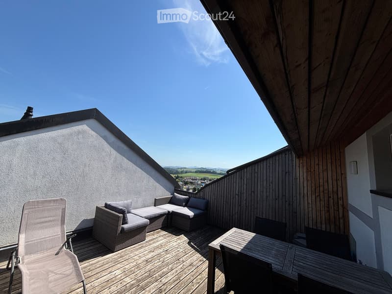 Wooden decked balcony with a grey wicker sofa, a wooden table, and chairs, overlooking a scenic countryside view.