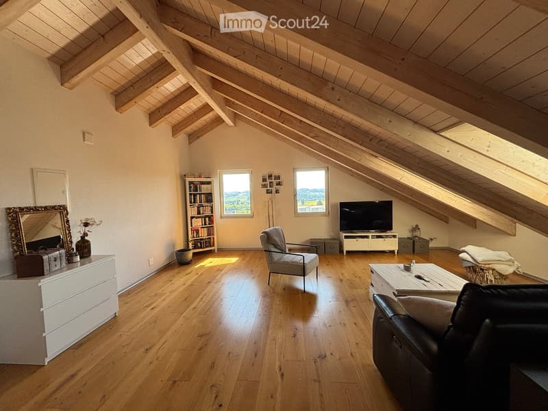 Bright living room with wooden floors, exposed wooden beams, large windows, sofa, armchair, coffee table, bookshelf, TV, and decorative items.