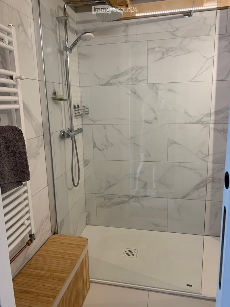 Modern bathroom with marble tiles, glass-enclosed shower, handheld showerhead, radiator, and wooden bench.