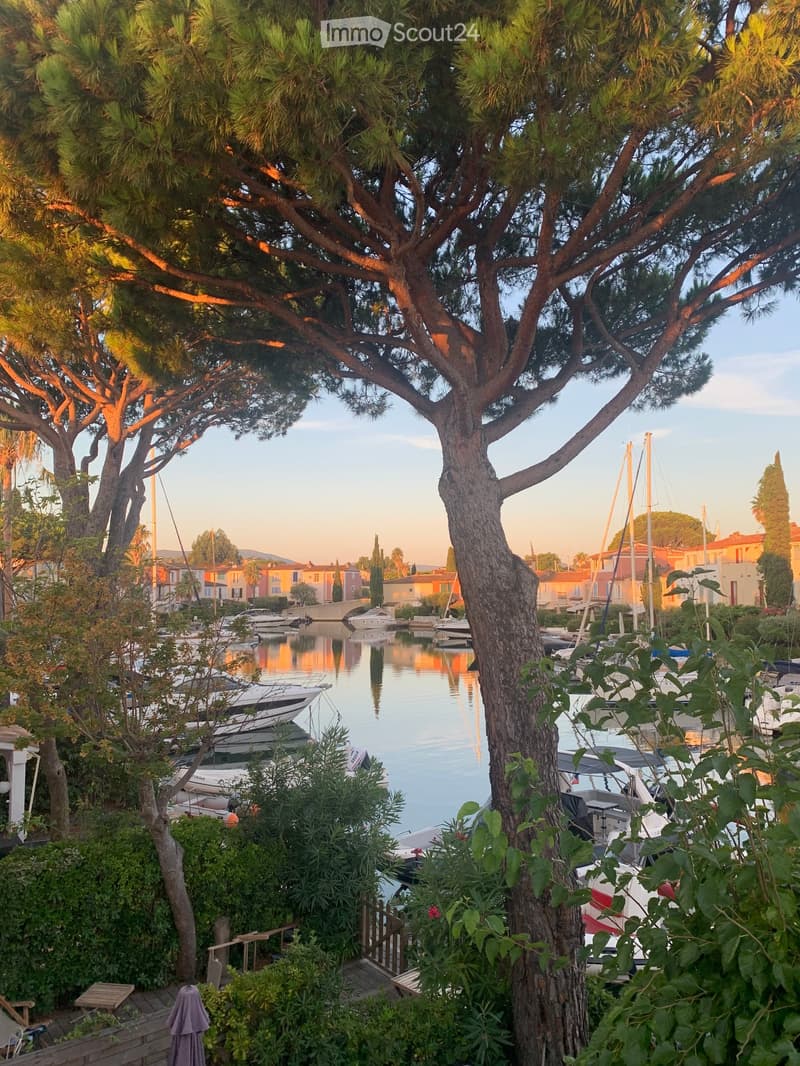A serene waterfront scene with a calm canal, several docked boats, and surrounding greenery, including trees and shrubs, with colorful houses in the background.