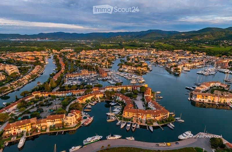 Aerial view of a coastal town with a marina filled with numerous boats and yachts, surrounded by colorful buildings and a scenic backdrop of hills and mountains.