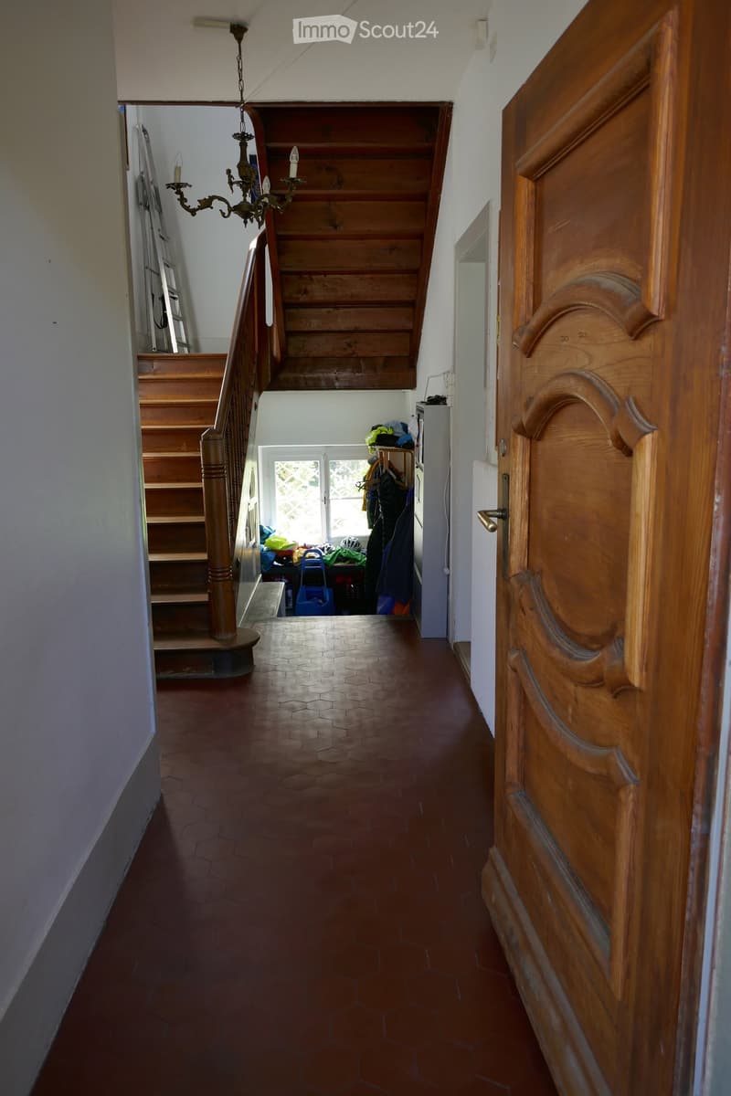 A narrow hallway with wooden stairs leading upwards, a chandelier hanging from the ceiling, and a wooden door on the right side. The floor is tiled in a hexagonal pattern, and the walls are painted white.