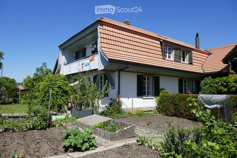 Two-story house with a red-tiled roof, white walls, and green shutters. Features a balcony on the upper floor with laundry hanging. The garden in front includes raised beds with vegetables and herbs, surrounded by shrubs and trees.